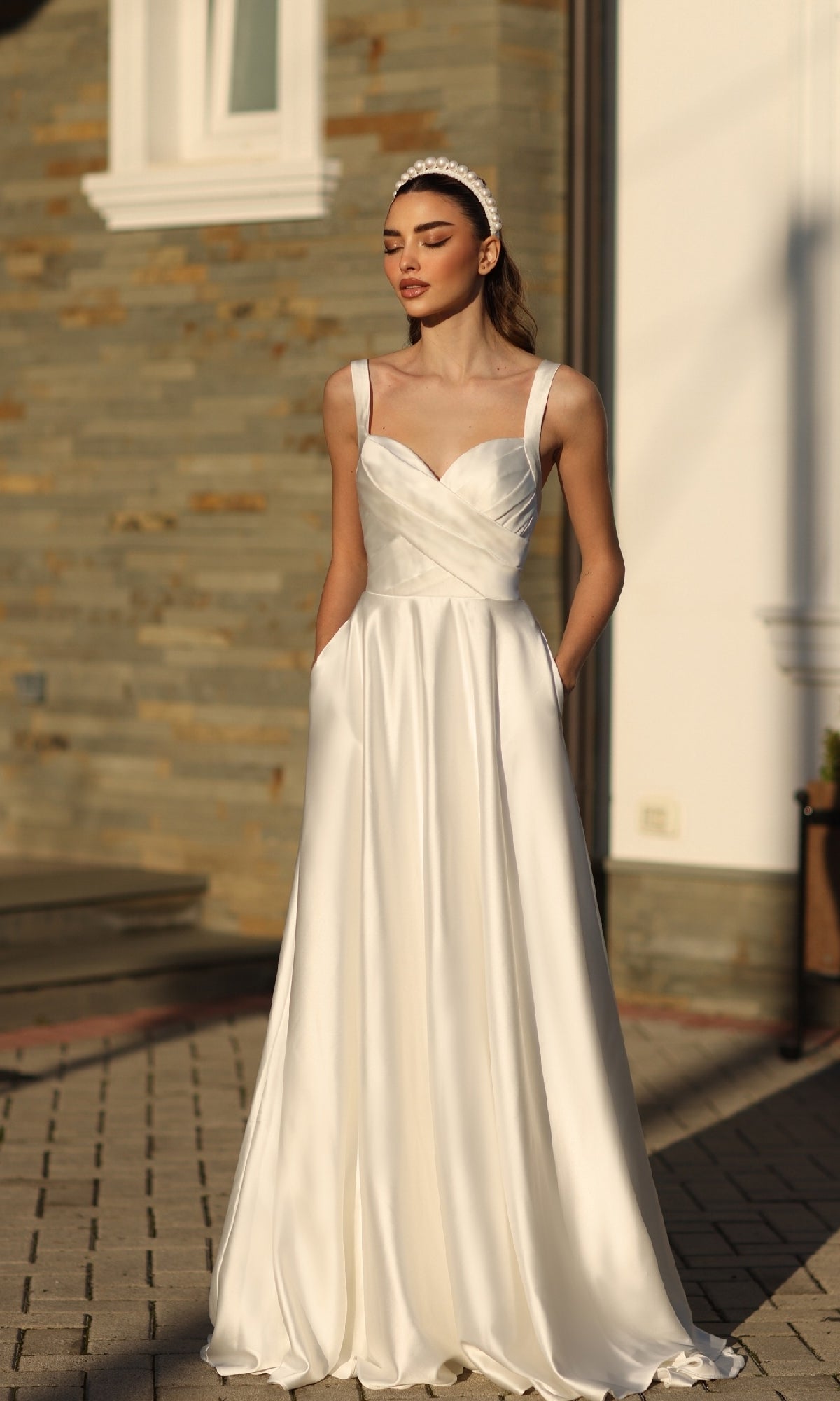 A woman stands outdoors in sunlight wearing the Off-White Satin Prom Ball Gown: Tina Holly BB210W, a sleeveless A-line dress featuring a fitted bodice and pockets.